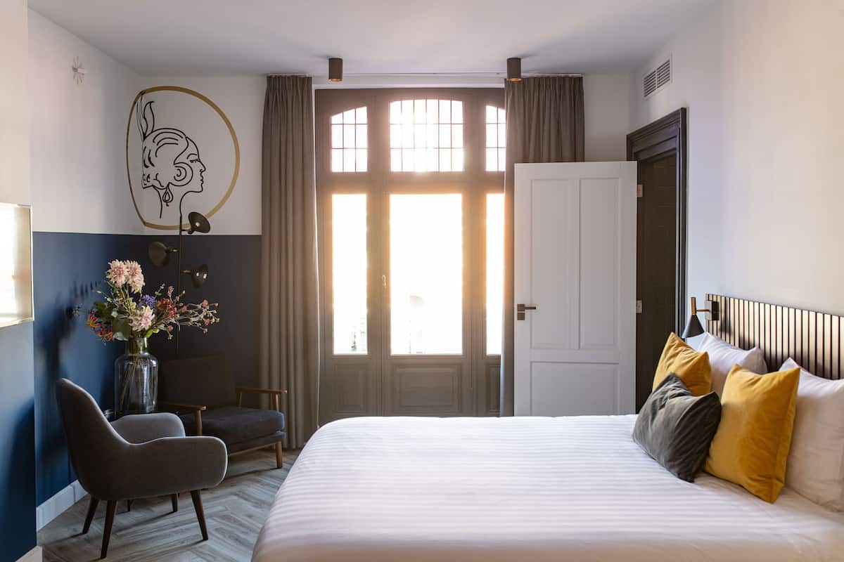 A hotel room with a large bed, a chair, a desk, a vase with flowers, and a window with curtains.