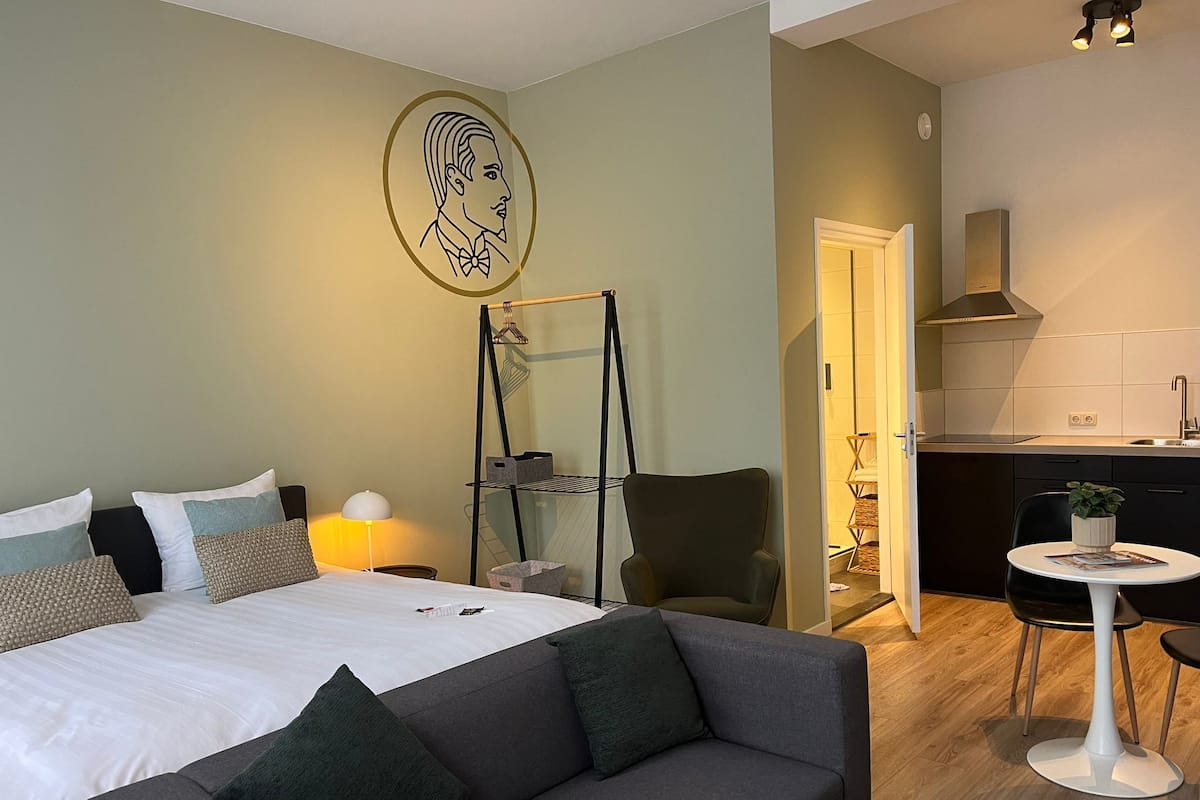 A modern hotel room with a bed, a grey sofa, a small round table, a chair, a kitchenette, and a wall-mounted shelf.