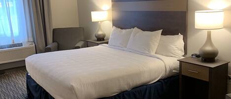 Una habitación de hotel con una cama grande, dos mesitas de noche con lámparas, una silla y un ventanal con cortinas.