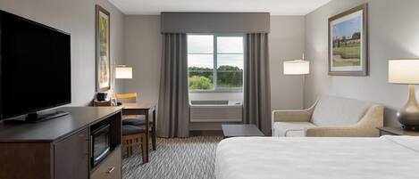 A hotel room with a bed, a desk with a chair, a television, a window with curtains, and a lamp.
