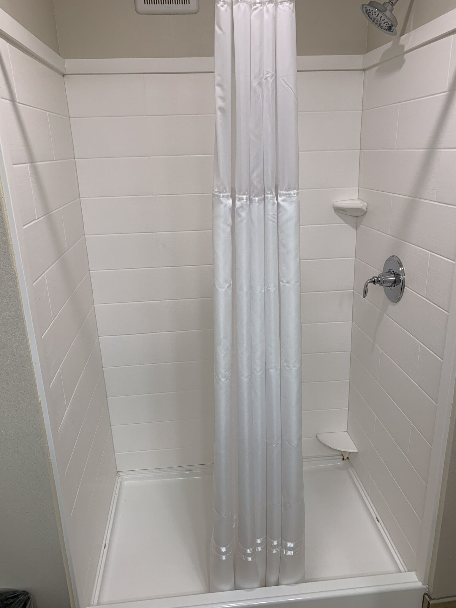 Une cabine de douche carrelée de carreaux blancs, avec un rideau de douche transparent, une douchette à main et une petite étagère blanche.