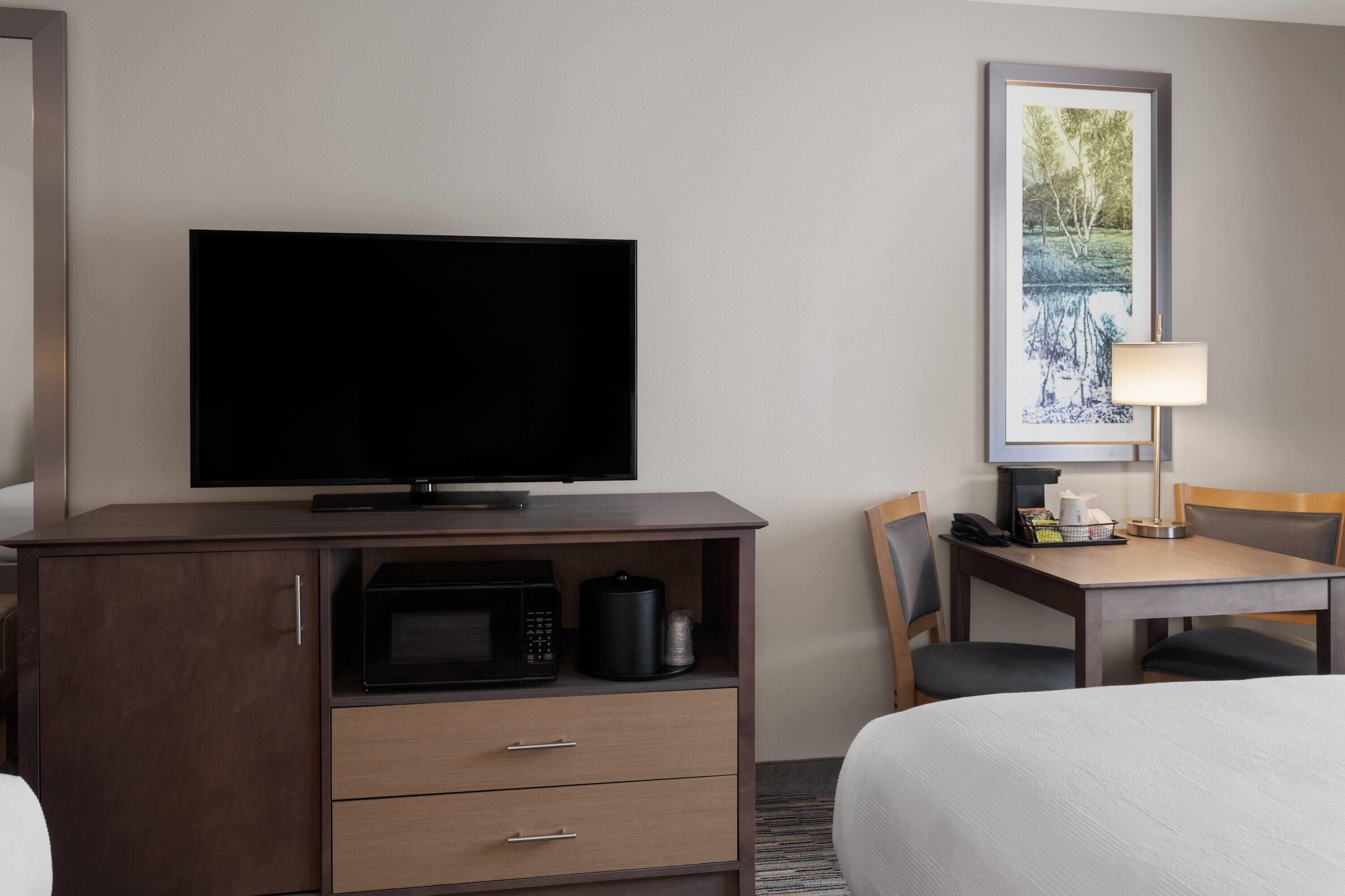 A hotel room with a flat-screen TV on a wooden cabinet, a microwave, a coffee maker, and a dining table with chairs.
