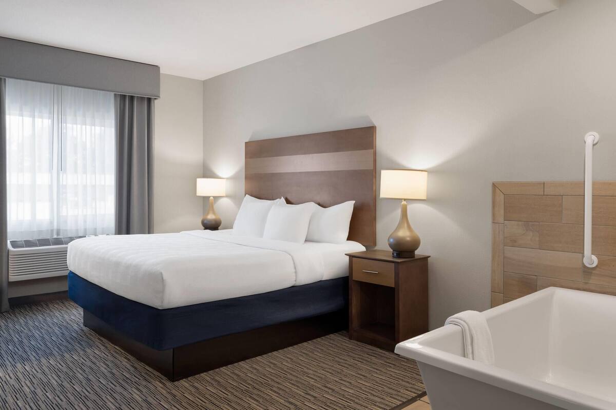 Habitación de hotel con cama, lámparas de noche, mesita de noche, bañera y ventana con cortinas.