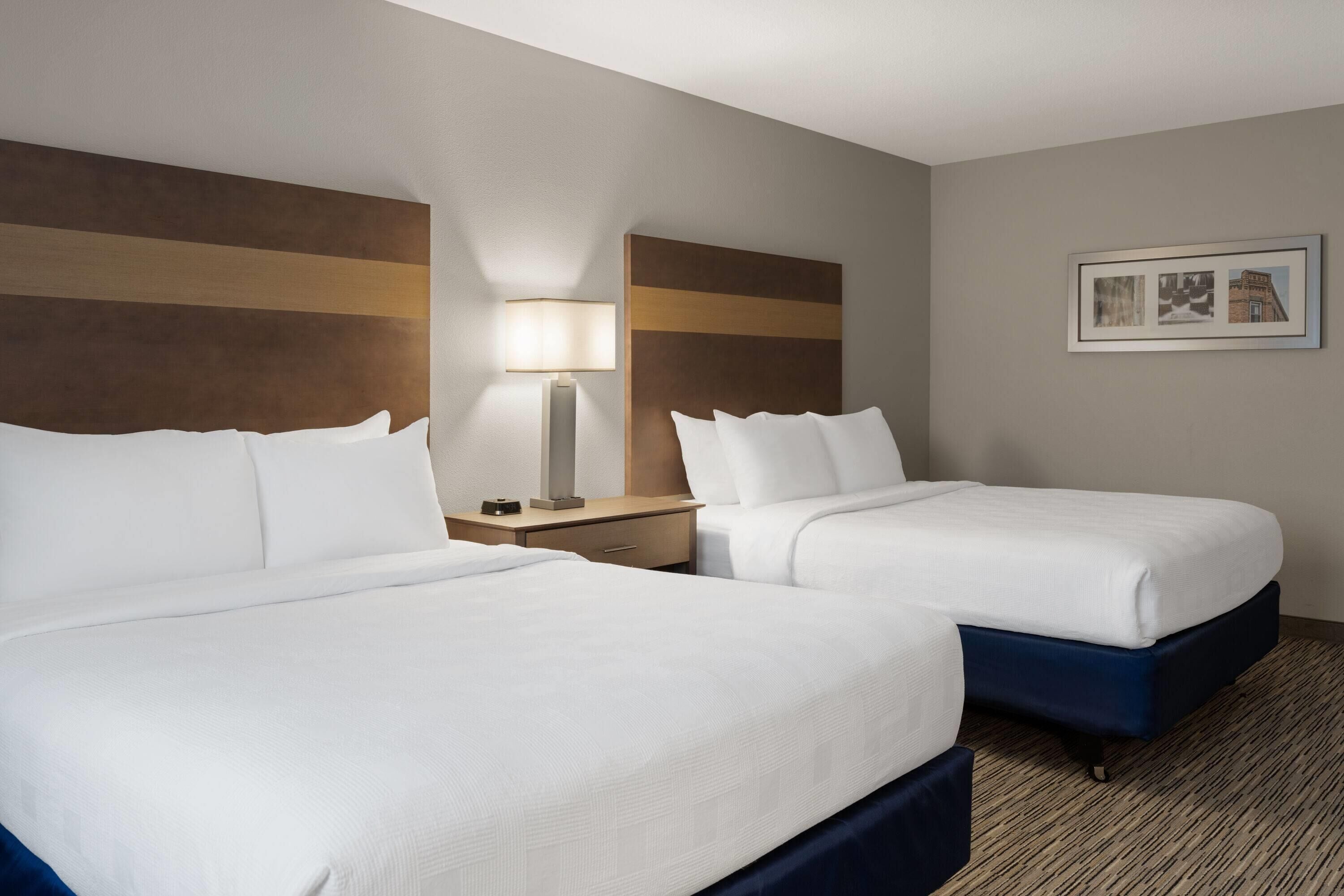 A hotel room with two beds, a nightstand, a lamp, and a framed picture on the wall.