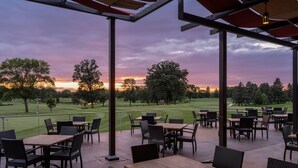 Un patio al aire libre con mesas y sillas, con vista a un campo de golf durante el atardecer.