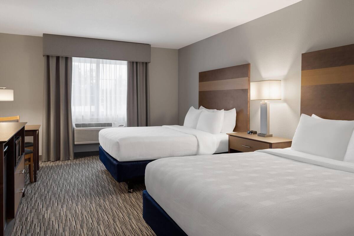 Habitación de hotel con dos camas, un escritorio, una lámpara y un ventanal con cortinas.
