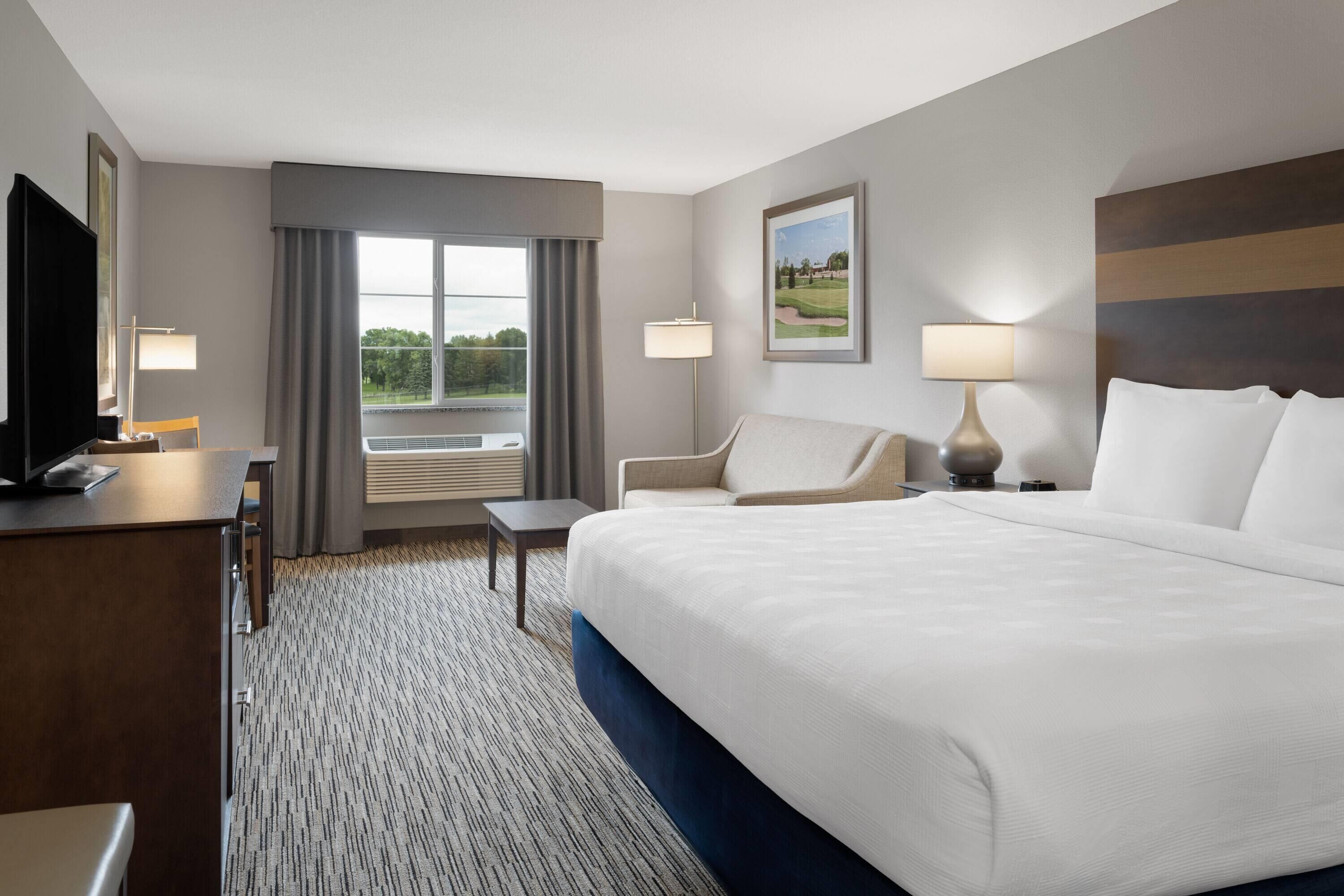 Habitación de hotel con una cama grande, un escritorio con silla, una mesita, dos lámparas y un ventanal con cortinas.