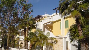 Un edificio con fachada amarilla, paredes blancas y contraventanas verdes. Se ven palmeras y una pérgola con vigas de madera.