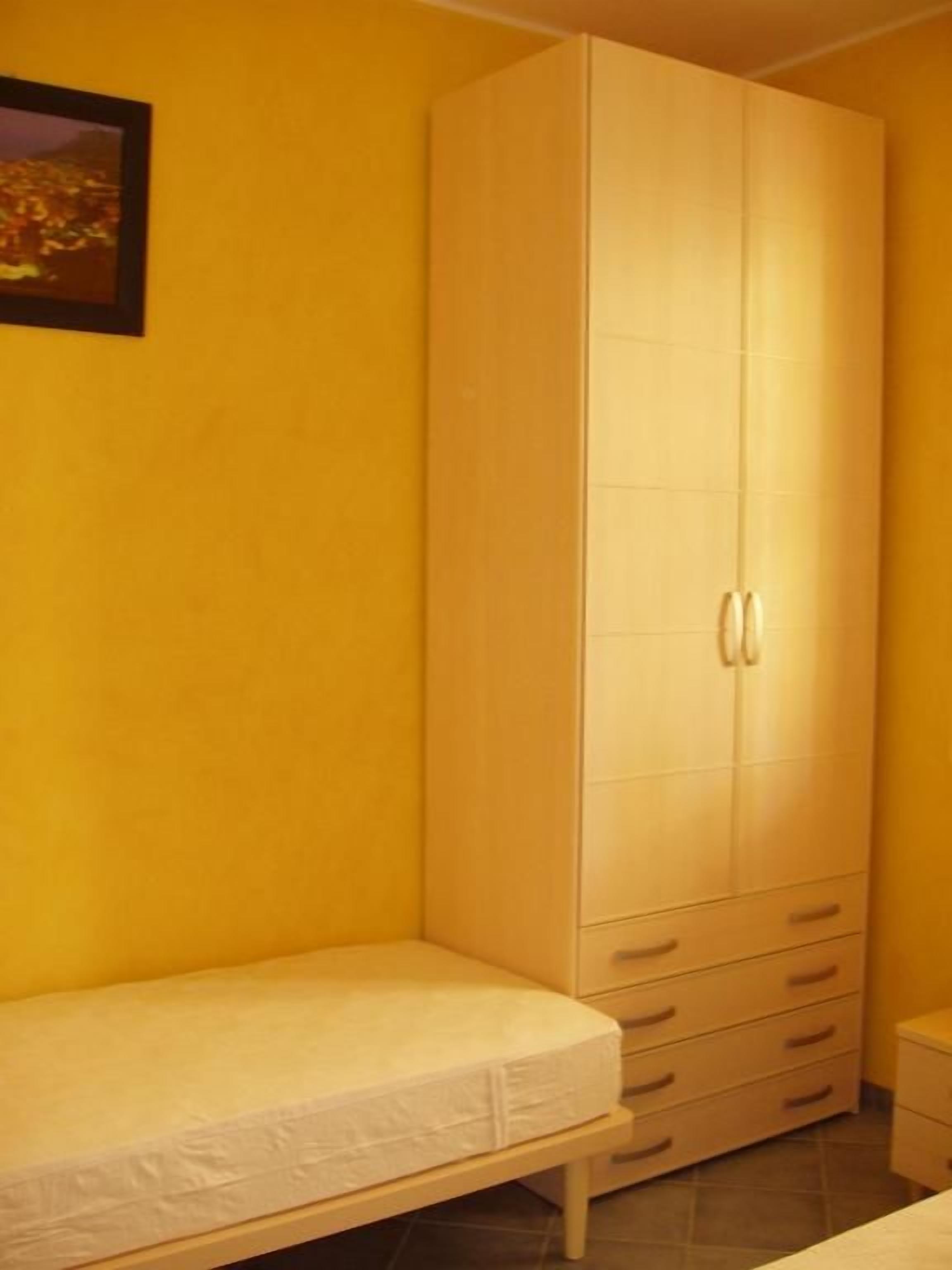 Ein kleines Zimmer mit einem Einzelbett, einem Kleiderschrank mit Schubladen und einem gerahmten Bild an der Wand.