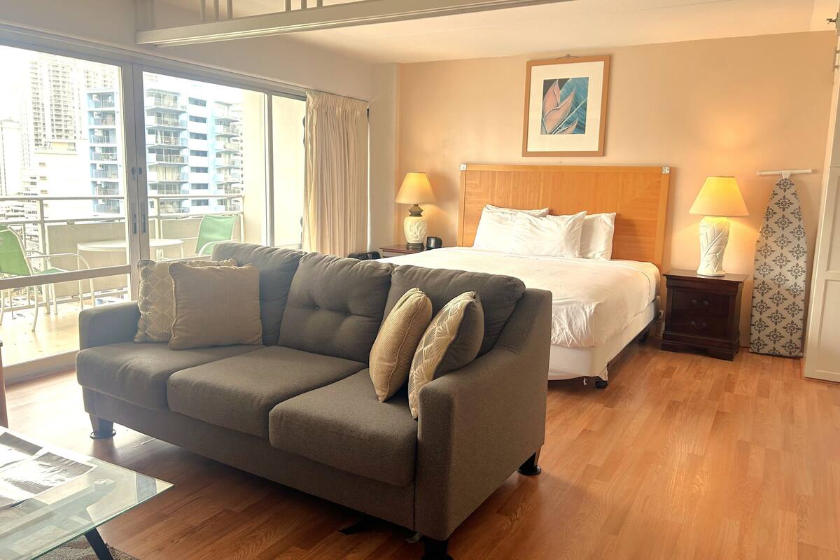 A hotel room with a sofa, a bed, a nightstand, a lamp, a window with curtains, and a painting on the wall.