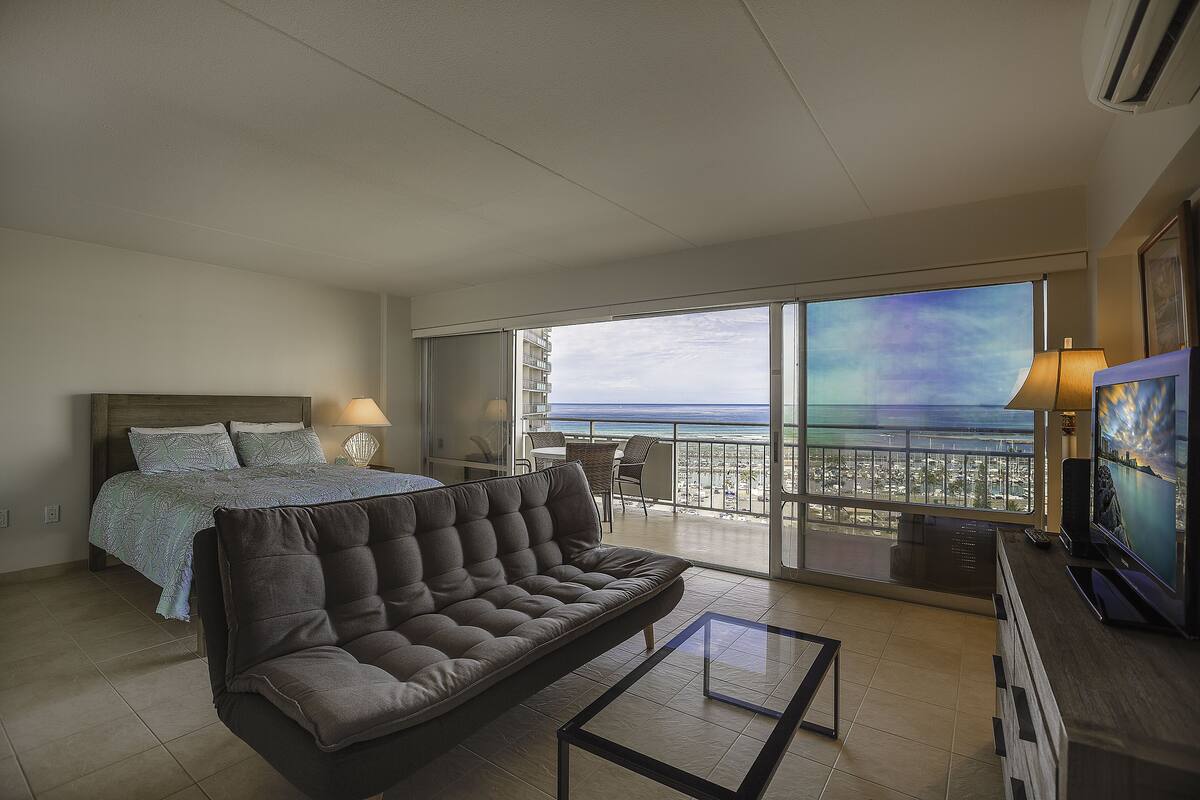 A modern hotel room with a bed, sofa, balcony with a view of the city, and a TV.