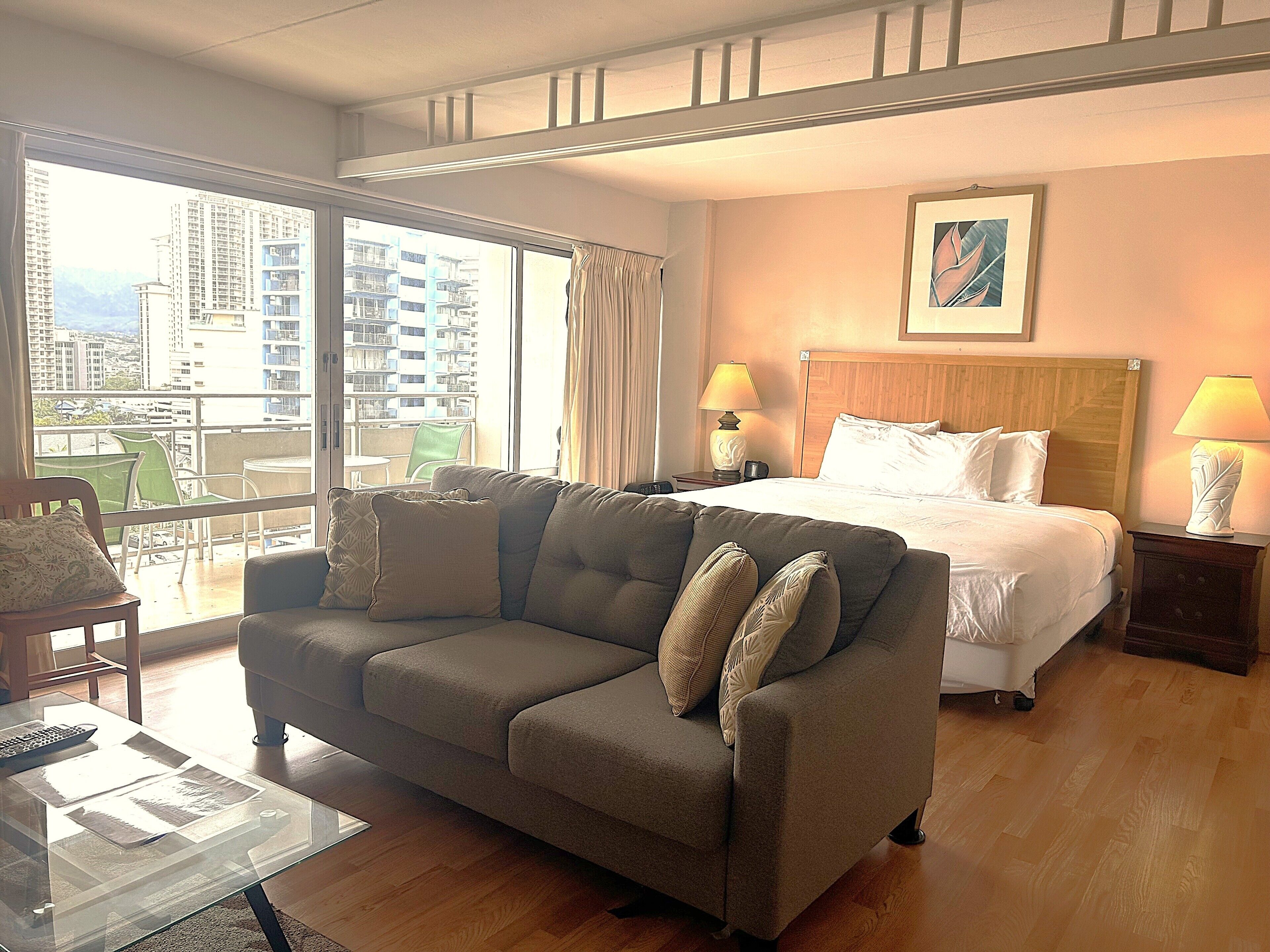 A hotel room with a large bed, a sofa, a balcony with a view of high-rise buildings, and two bedside lamps.