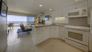 A modern kitchen with white cabinets, a built-in microwave, and an oven. There is a balcony with a chair and a table, and a view of the ocean. - Ilikai by Luana Hawaii Vacation Rentals (Honolulu)