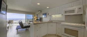 A modern kitchen with white cabinets, a built-in microwave, and an oven. There is a balcony with a chair and a table, and a view of the ocean.