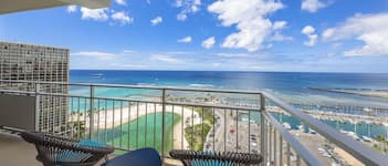 A balcony with wicker chairs and a table, overlooking a beach and marina with boats.