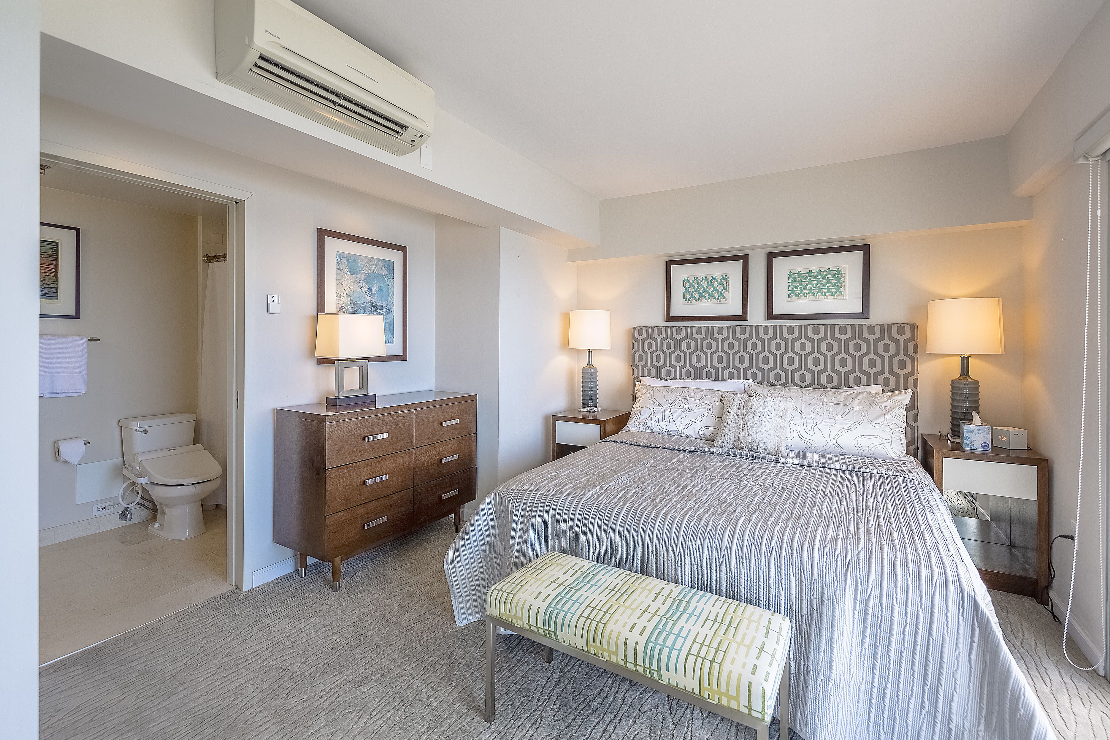A bedroom with a bed, bedside tables, a bench, a dresser, a nightstand, and a wall-mounted air conditioner.