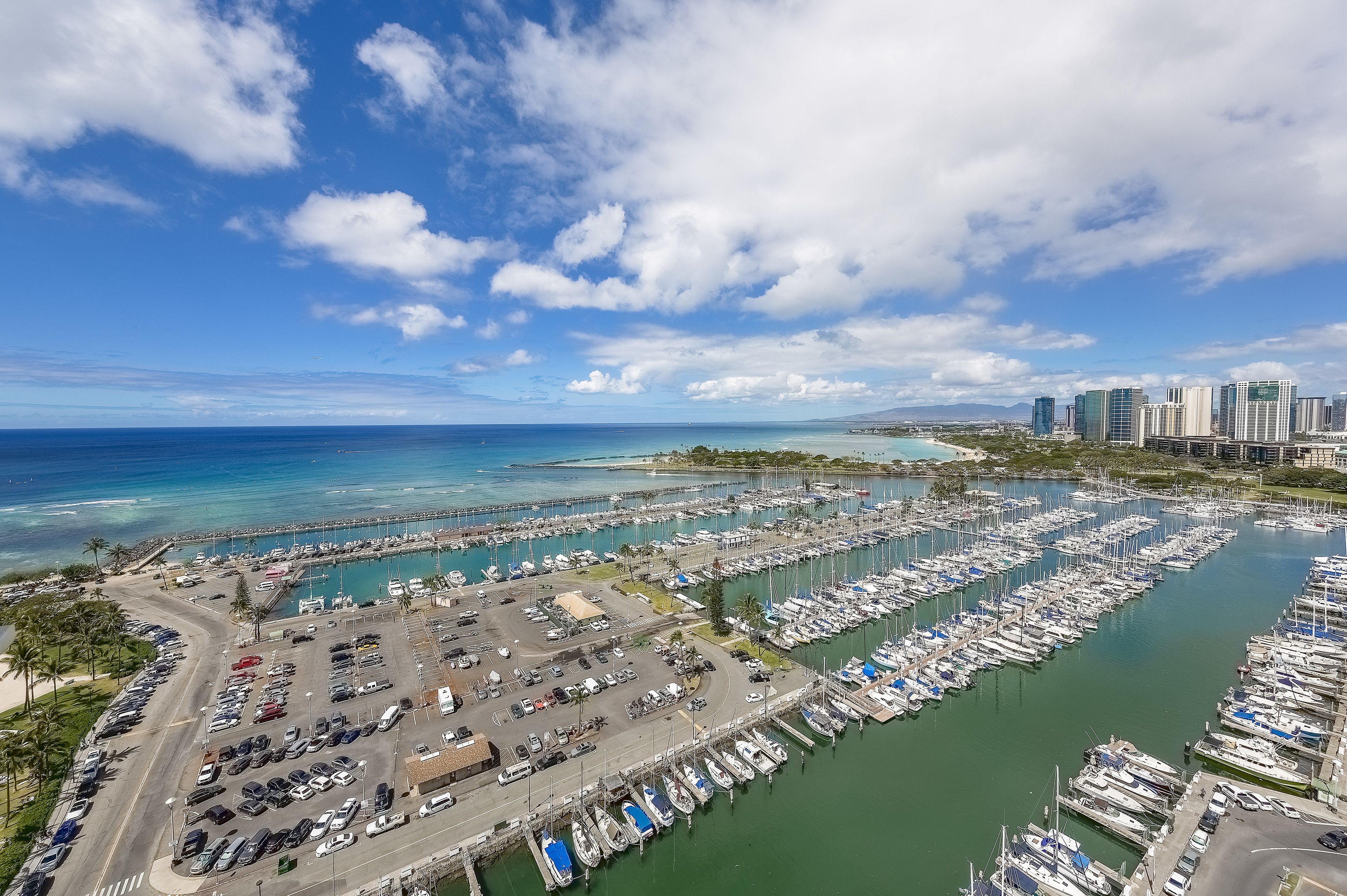 An aerial view of a marina with numerous boats docked, a parking area, and a cityscape in the background.