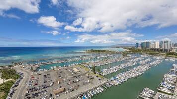 An aerial view of a marina with numerous boats docked, a parking area, and a cityscape in the background.