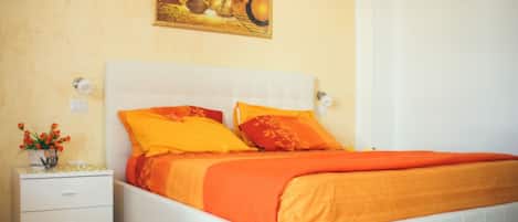 A bedroom with a white bed, orange bedding, a nightstand with a vase of flowers, and a painting on the wall.