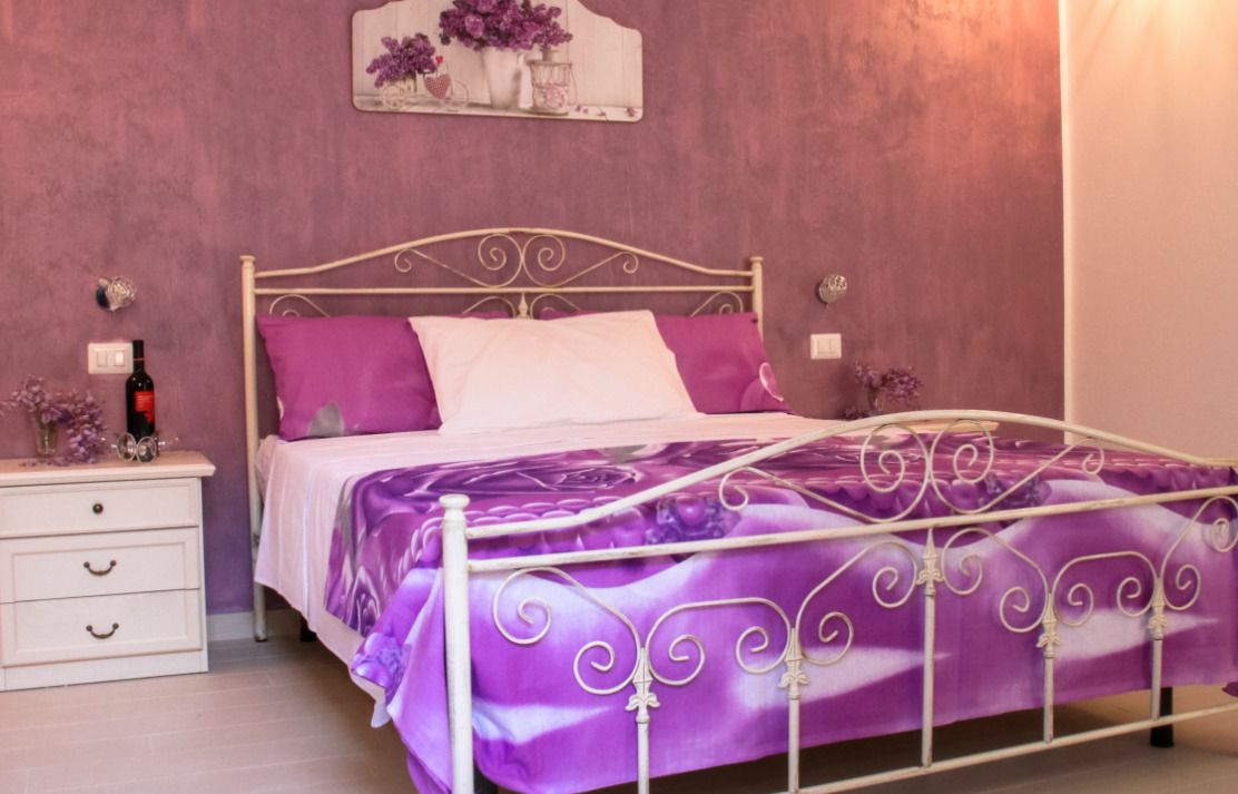 A bed with a purple bedspread and white headboard, a white nightstand with a bottle and a decorative item, and a wall-mounted mirror with purple flowers.