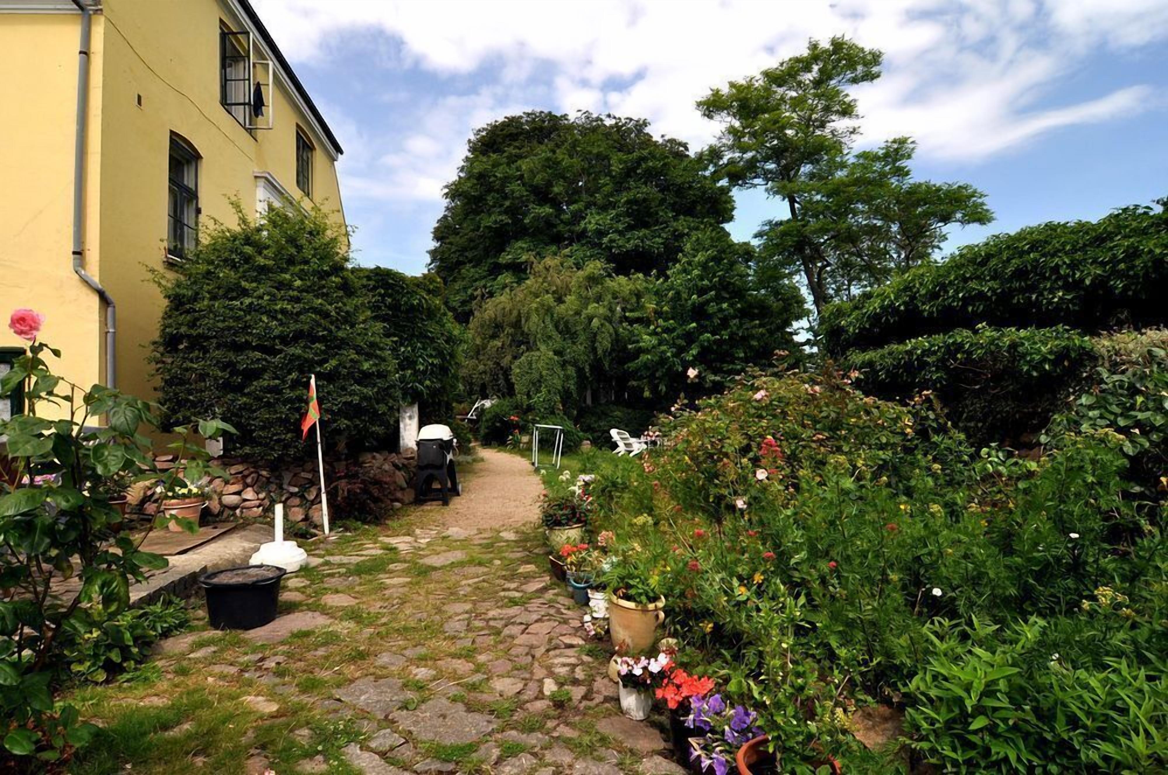 Un sendero de jardín que conduce a un edificio con fachada amarilla, rodeado de macetas con plantas y flores diversas.