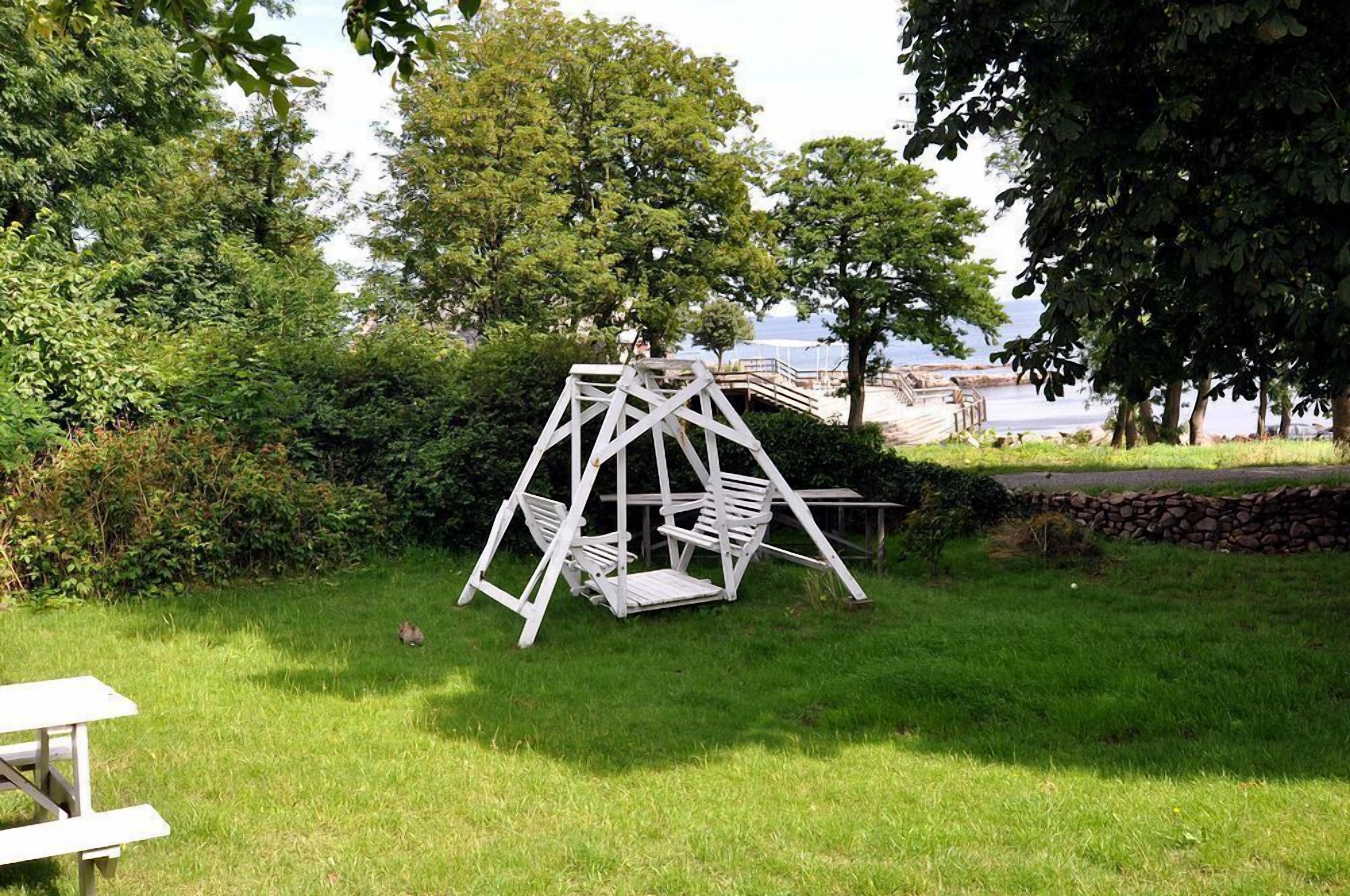 A white swing set in a grassy area with trees and a stone wall, near a body of water and a wooden dock.
