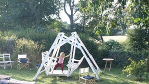 A child playing on a white swing set in a grassy outdoor area with trees and a bench in the background. - Danchelshus (Allinge)