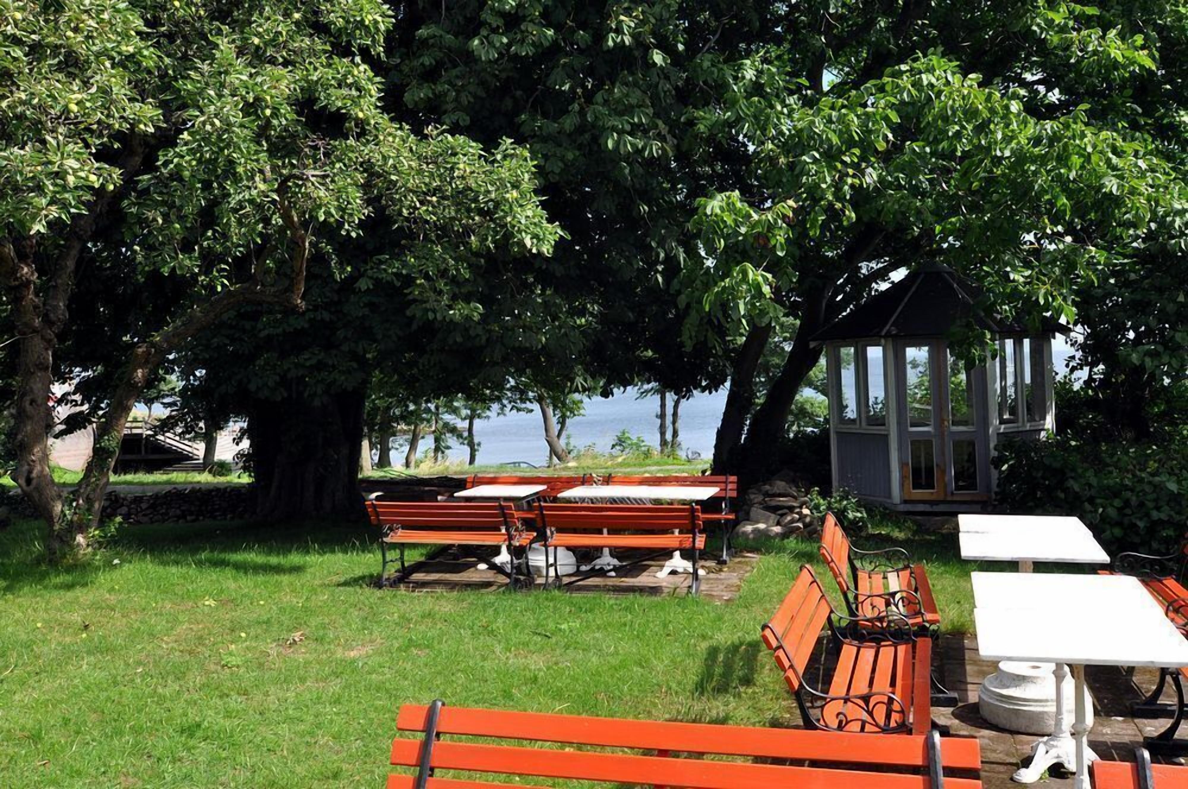 A park with benches and tables under large trees, a gazebo, and a view of the water.