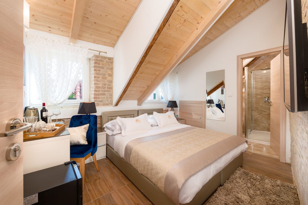 A bedroom with a large bed, a desk with a chair, a mirror, and a shower area.