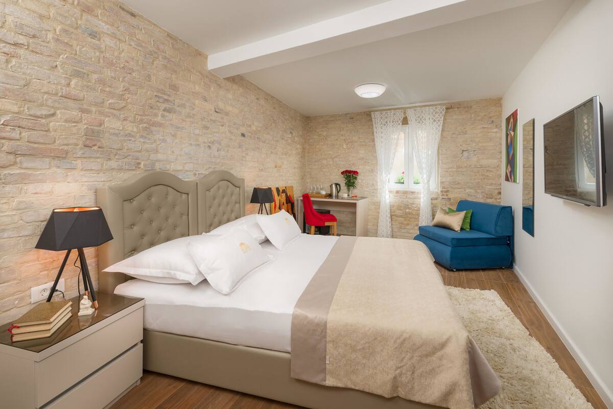 A bedroom with a brick wall, a bed with a beige headboard, a nightstand with a lamp, a desk with a chair, and a blue sofa.