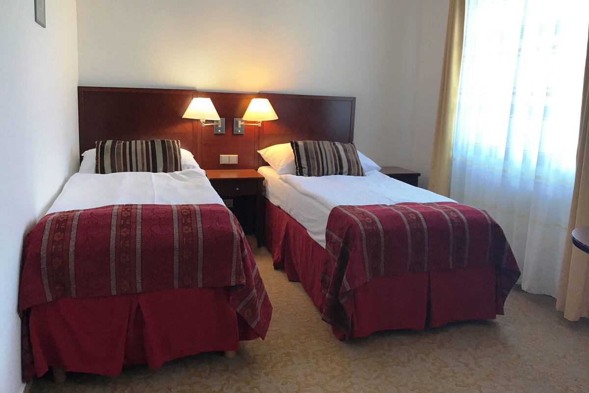 A hotel room with two single beds, each with a red bedspread, a wooden headboard, and two wall-mounted lamps.