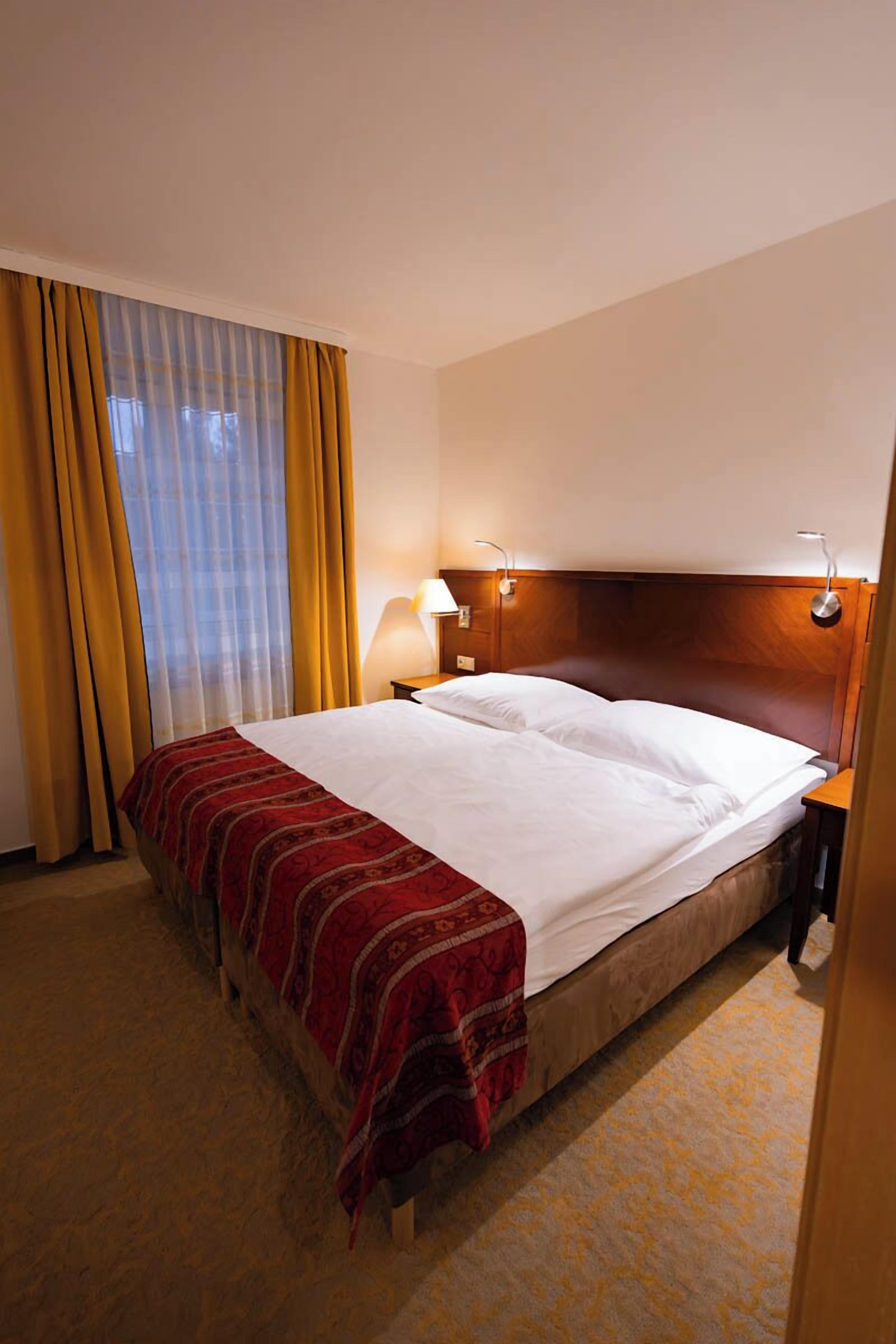 Una habitación de hotel con cama, mesita de noche, lámpara y ventana con cortinas.