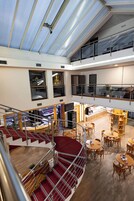 A spacious hotel lobby with a central staircase, multiple dining areas, and a mezzanine level with a glass railing.