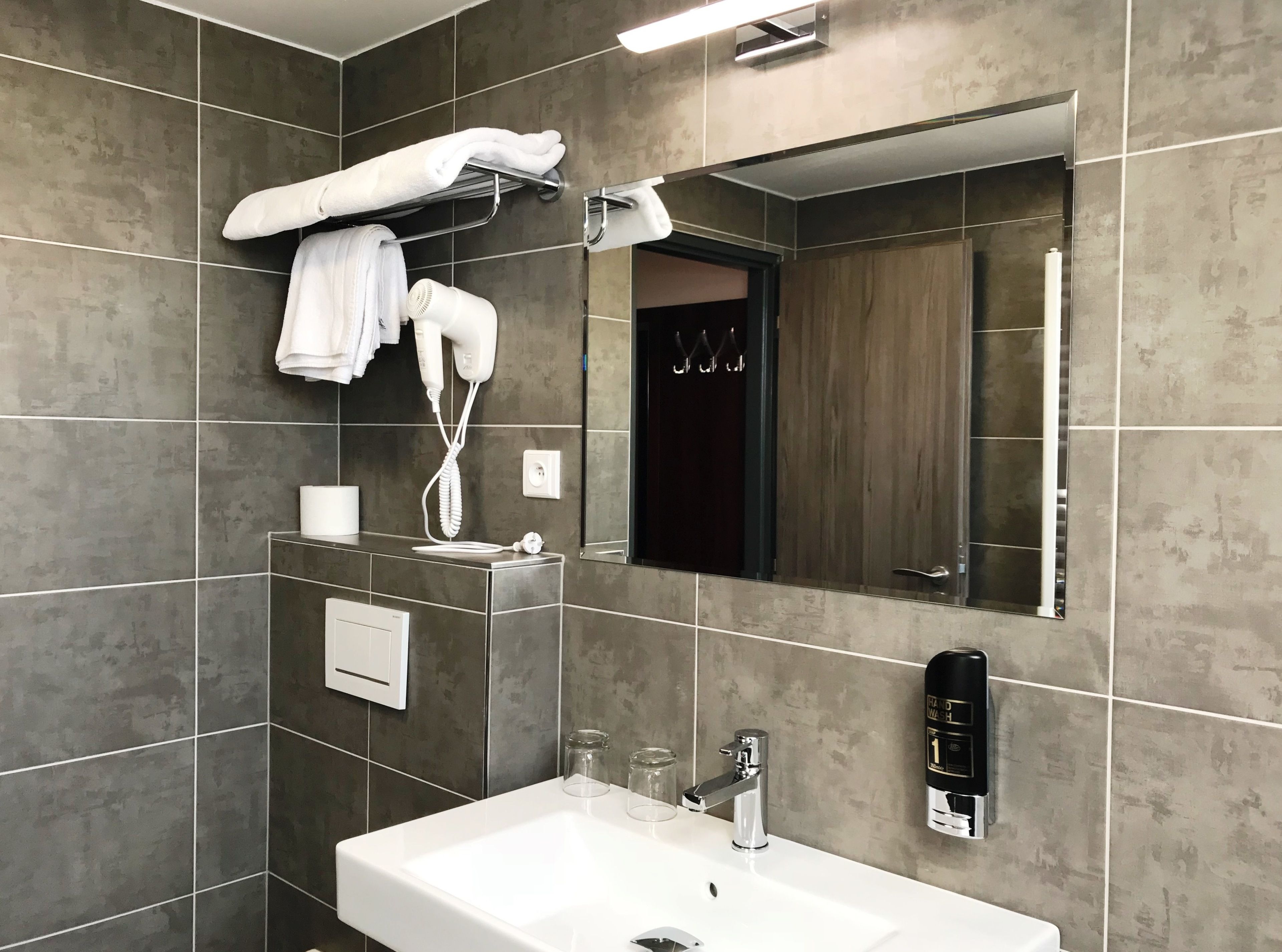 A modern bathroom with a large mirror, a white sink, a towel rack, a hairdryer, and a soap dispenser.