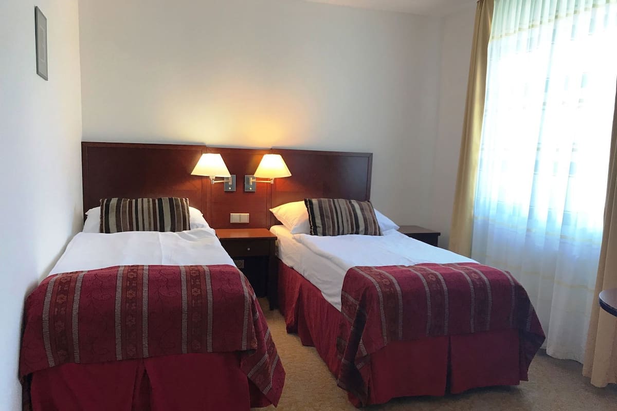 A hotel room with two beds, each with a red bedspread, a wooden headboard, two wall-mounted lamps, and a window with curtains.