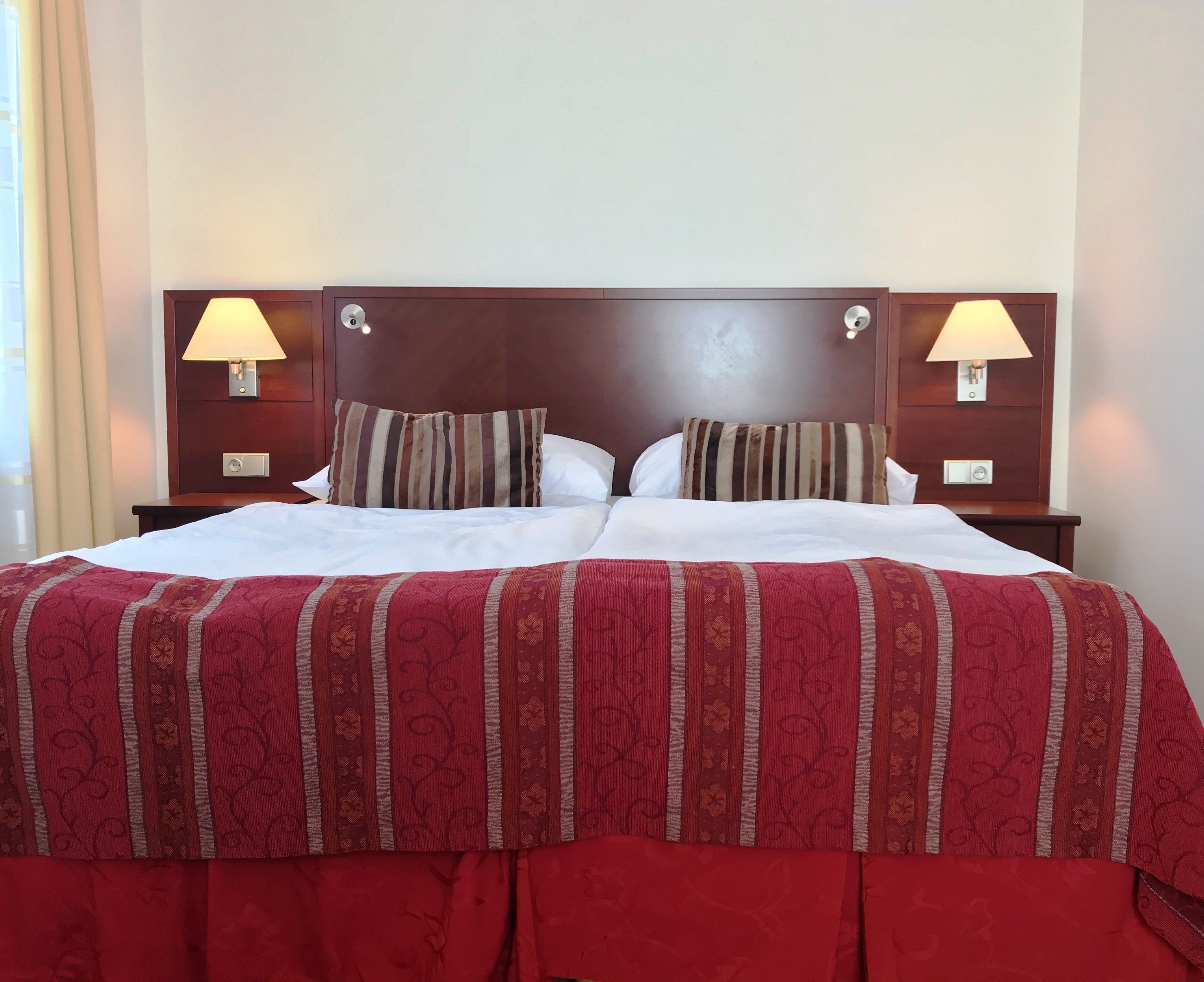 Un lit double avec une couvre-lit à motifs rouges et beiges, deux lampes de chevet et deux coussins à rayures.