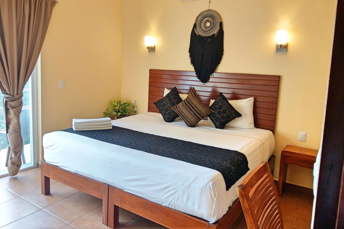 A hotel room with a wooden bed, white linens, a black throw, and decorative pillows. There is an air conditioning unit above the bed, a wall-mounted light fixture, and a small bedside table with a plant.