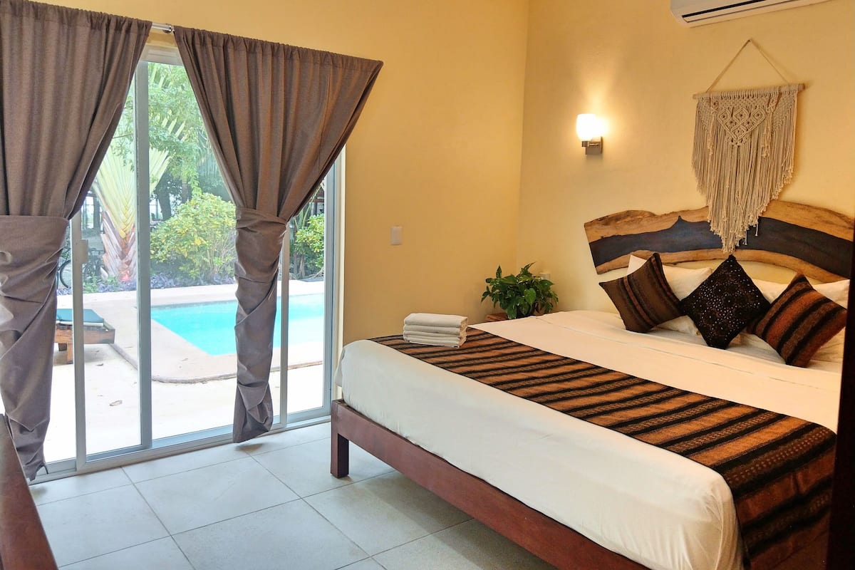 A bedroom with a bed, curtains, a wall-mounted air conditioner, a macrame wall hanging, and a view of a swimming pool.