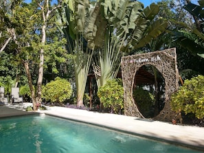 Un espace aménagé au bord de la piscine, avec une eau cristalline, une végétation luxuriante et une arche décorative portant l’inscription « Tulum Jah ».