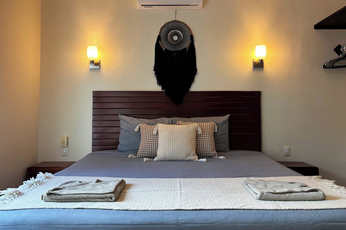A neatly made bed with a dark headboard, two wall-mounted lights, and a wall-mounted air conditioner.