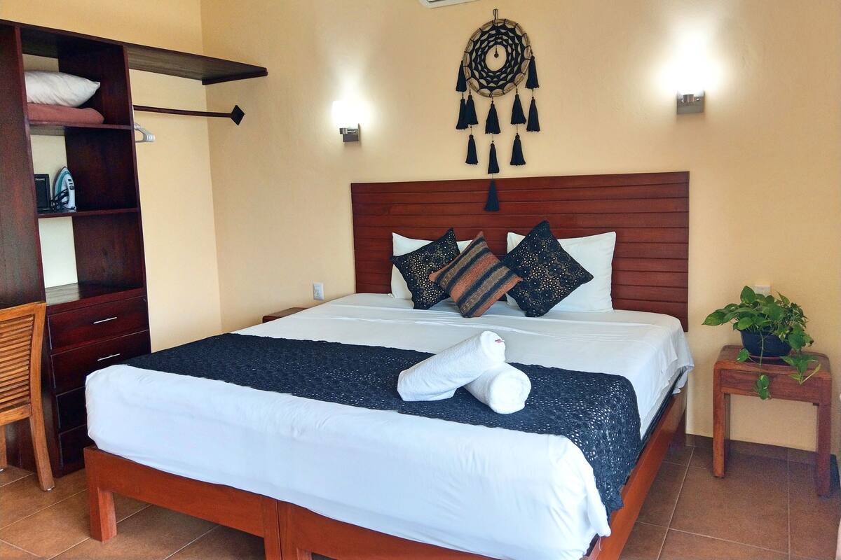 A neatly made bed with white linens and a dark blue blanket, a wooden headboard, a wall-mounted air conditioner, a decorative wall hanging, and a small wooden nightstand with a plant.