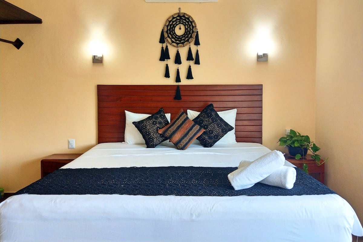 A neatly made bed with white linens and multiple decorative pillows, a wooden headboard, a wall-mounted air conditioner, and a wall hanging.