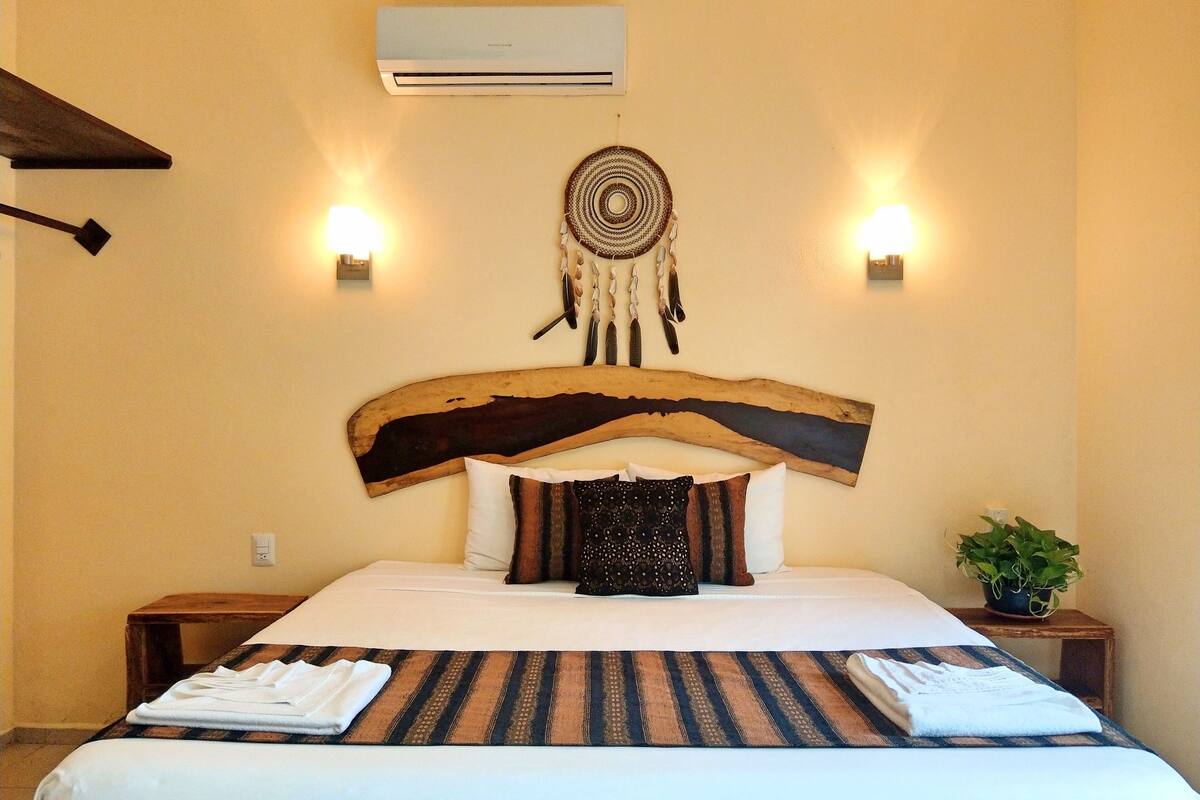 A hotel room with a large bed, a wooden headboard, two wall-mounted lights, a wall-mounted air conditioner, and a small potted plant.