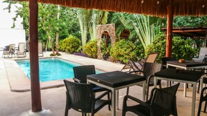 An outdoor pool area with lounge chairs and tables, surrounded by lush greenery and a thatched roof structure. - Tulum Nah (Tulum)