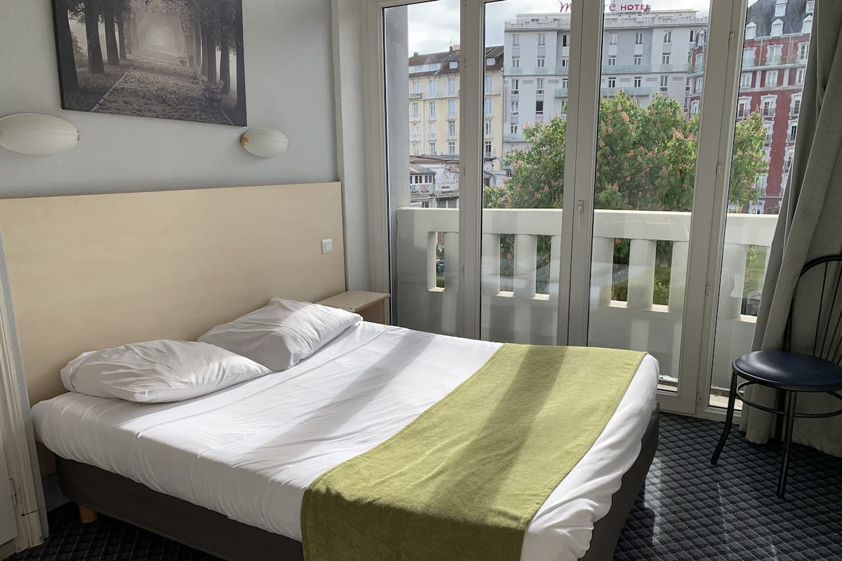 Een hotelkamer met een bed, een groene plaid, een stoel en uitzicht op gebouwen door het raam.
