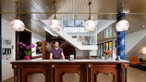 A reception desk with a man standing behind it, a staircase in the background, and a blue column. - Hôtel Hélianthe (Lourdes)