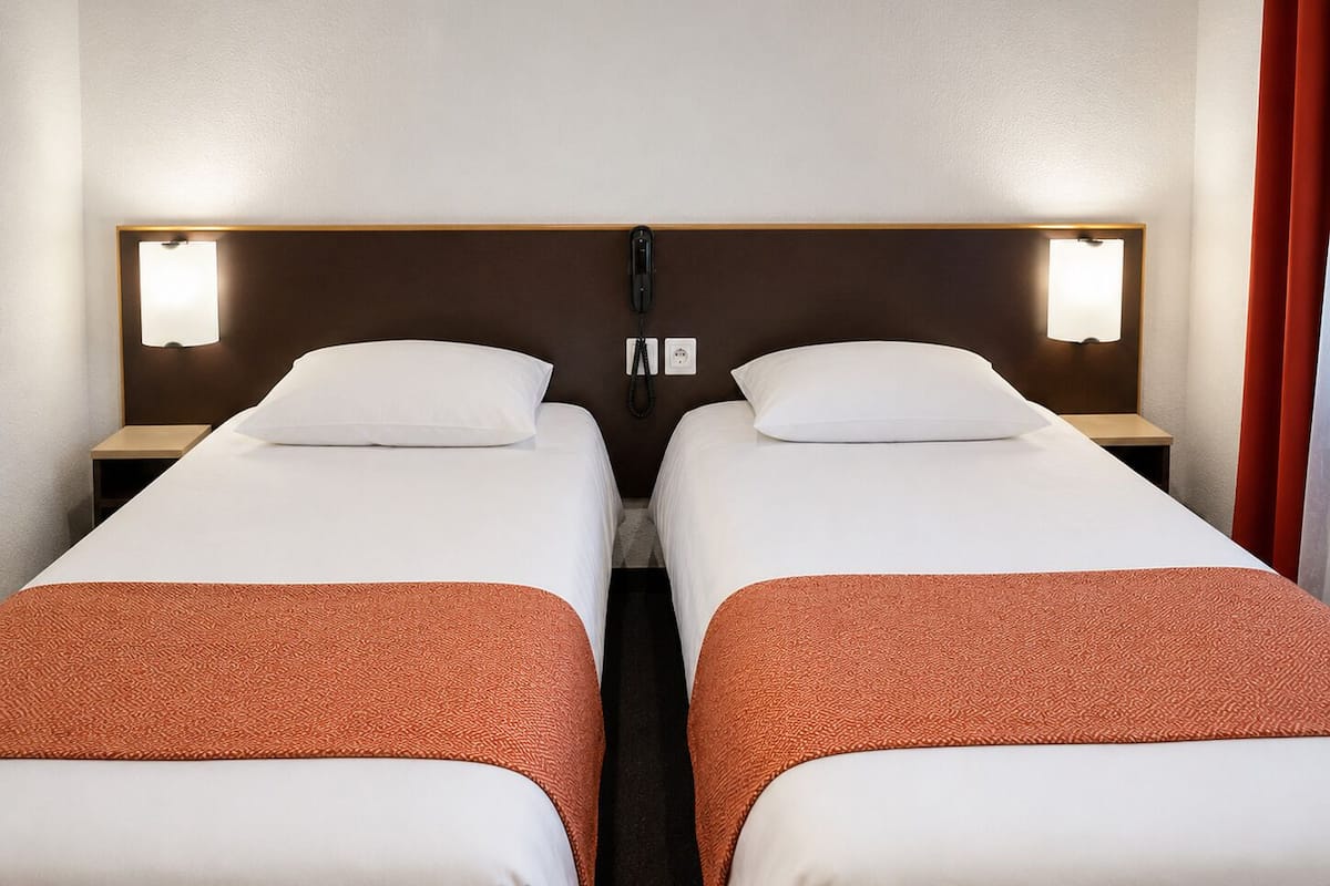 A hotel room with two single beds, each with a pillow and a blanket. There are bedside tables with lamps and a phone on each.