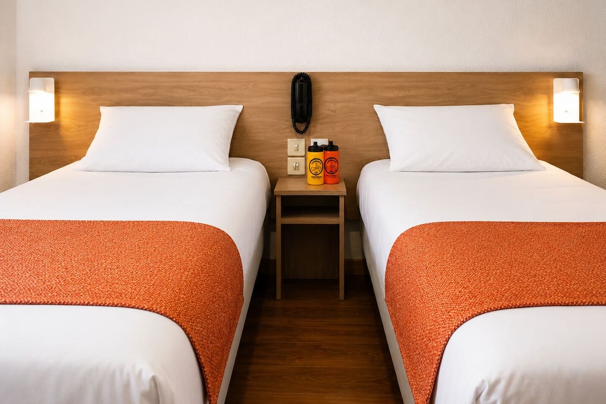 A hotel room with two beds, each with white bedding and orange throw pillows. There is a wooden nightstand between the beds with two bottles and a phone.