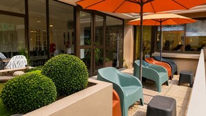 An outdoor seating area with chairs and tables, a large orange umbrella, and a glass door entrance. - Hôtel Hélianthe (Lourdes)