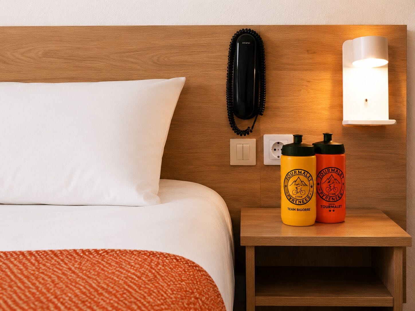 Una habitación de hotel con cabecera de madera, almohada blanca, lámpara de noche, teléfono y dos botellas de agua con el logo de la marca sobre una mesita de noche de madera.